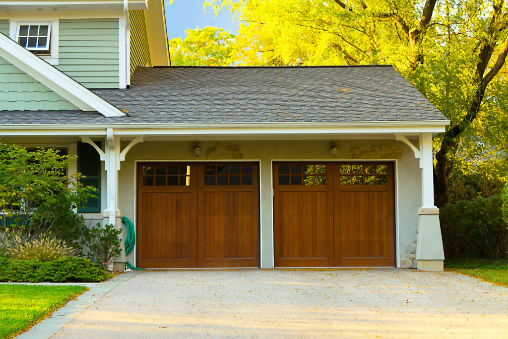 Two wood garage doors with small windows in the top, in a light green home with trees in background.