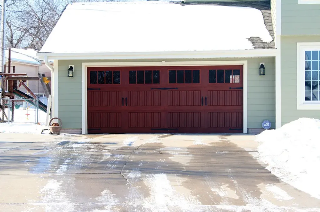 Dark red garage door with snowy driveway and snow on roof. Home with light green siding.